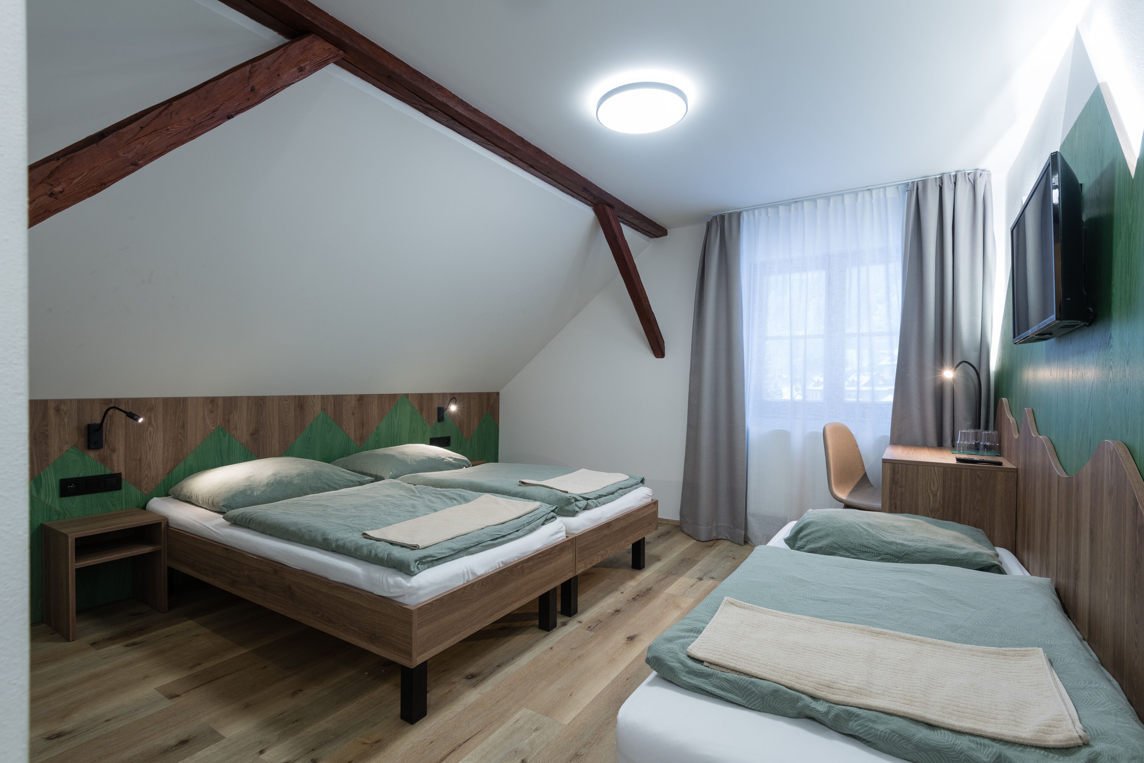 A room with two wooden beds, a desk with a chair, a TV mounted on the wall, and a window with curtains.