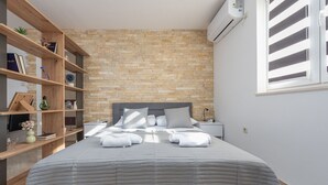 A bedroom with a brick wall, a bed with a grey bedspread, a wooden bookshelf, and a window with blinds. - Villa Izvor with heated pool, rural place (Bribir)