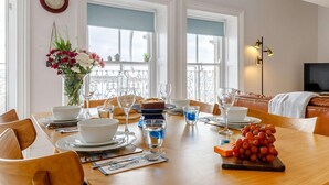 Zona pranzo con tavolo di legno apparecchiato per quattro persone, circondato da sedie. Sul tavolo c'è un vaso con fiori e un orologio appeso al muro.