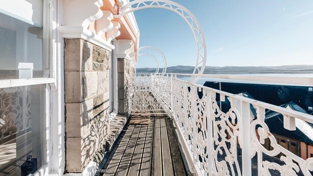 Un balcone con ringhiere bianche decorate, pavimento in legno e vista libera sul mare e sulle montagne in lontananza.