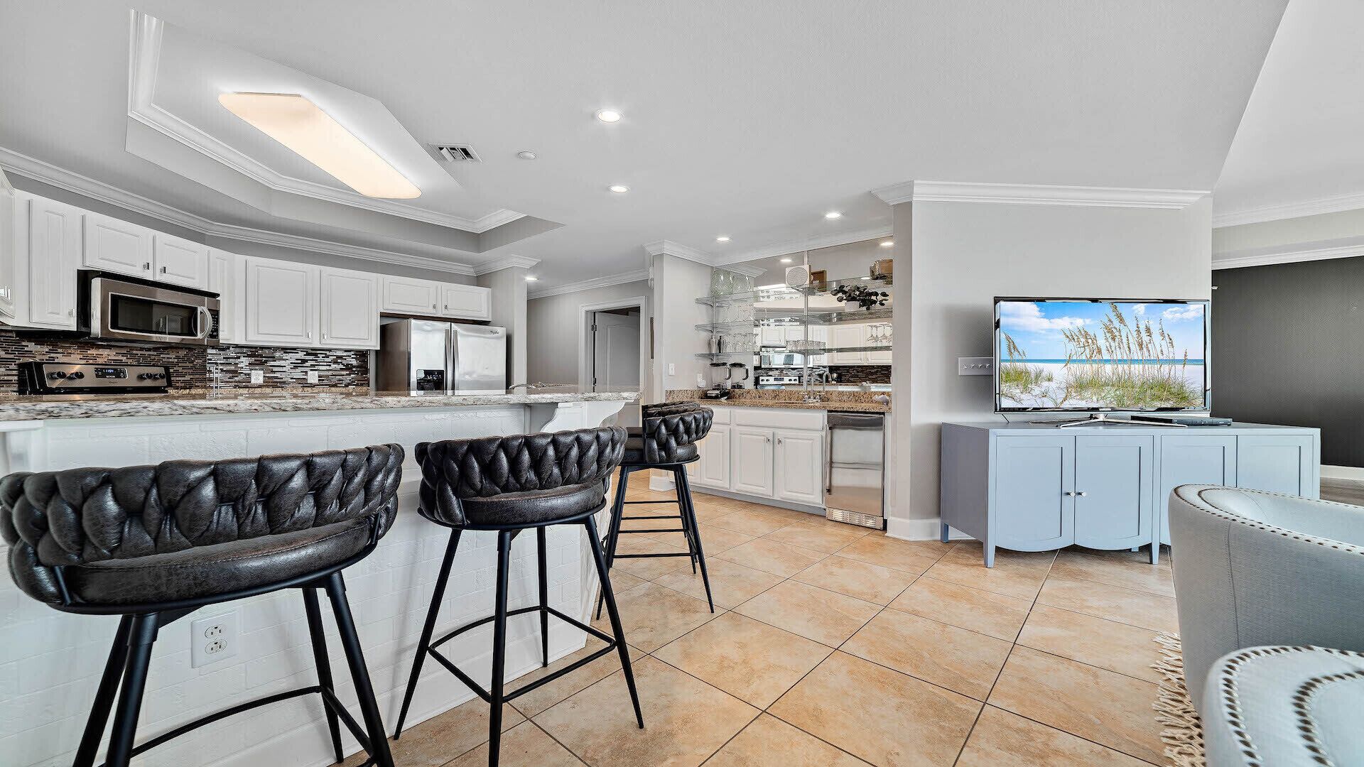 A modern kitchen with white cabinetry, a central island with bar stools, a built-in oven, and a large flat-screen TV on a cabinet.