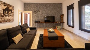Uma sala de estar moderna com um sofá escuro, uma mesa de centro de madeira, uma televisão montada e uma janela grande.
