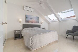 Relax in this serene bedroom with a large bed, bedside table, and chair, surrounded by natural light and a beautiful framed picture.