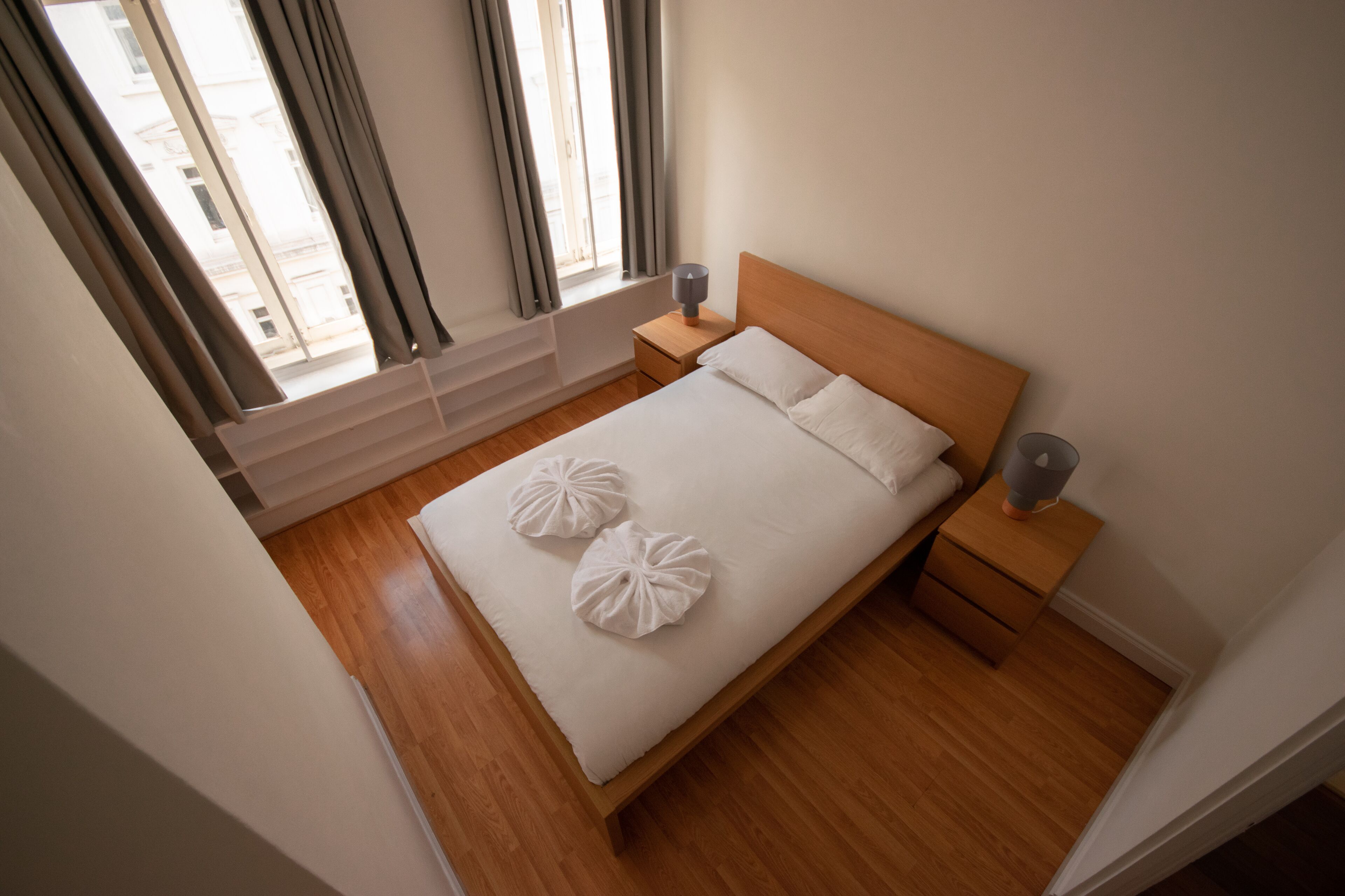 Una cama bien hecha con dos almohadones decorativos, un cabecero de madera, una mesita de noche con lámpara y una ventana con cortinas.