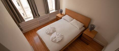 Una cama bien hecha con dos almohadones decorativos, un cabecero de madera, una mesita de noche con lámpara y una ventana con cortinas.