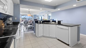 Une cuisine moderne avec des meubles blancs, un plan de travail noir et un lave-vaisselle. On aperçoit, au fond, un coin repas avec une table et des chaises.