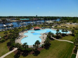 A large, irregularly shaped swimming pool surrounded by palm trees and lounge chairs, with a marina and boats in the background. - Yacht Club Villas #2-301 Amazing Views in Spacious End Unit! (North Myrtle Beach)