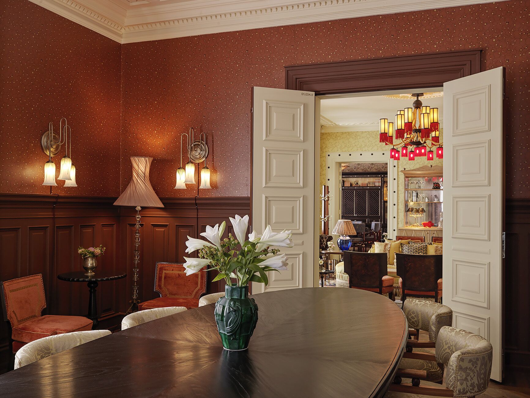 A formal dining room with a round table, chairs, a vase with flowers, wall sconces, and a chandelier.