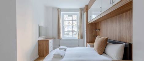 A modern bedroom with a large bed, wooden headboard, a bedside table, a TV, and a window with curtains.
