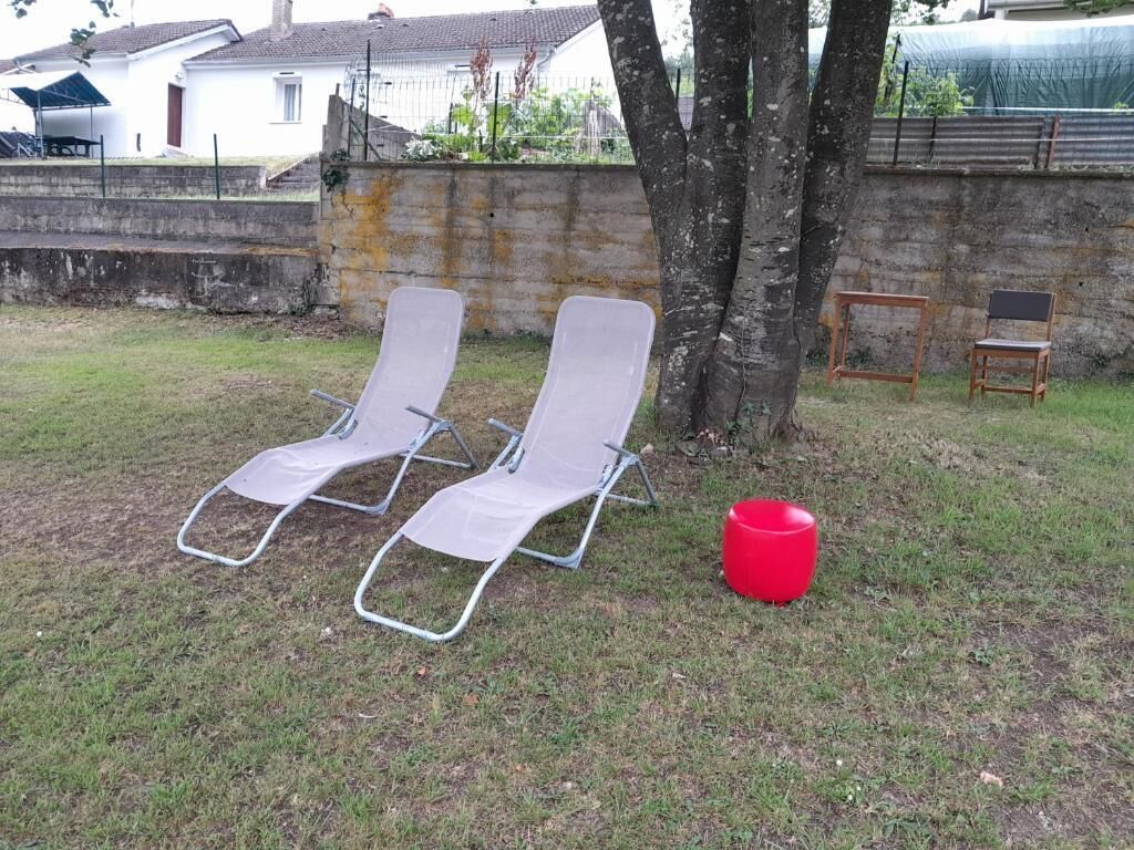 Duas cadeiras de lounge com estrutura de metal e assentos de tela, um banquinho de plástico vermelho, e uma árvore em área gramada com um muro de concreto ao fundo.