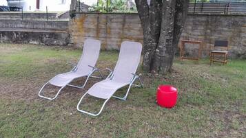 Duas cadeiras de lounge com estrutura de metal e assentos de tela, um banquinho de plástico vermelho, e uma árvore em área gramada com um muro de concreto ao fundo.