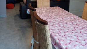 A dining area with a table covered in a floral tablecloth, a wicker chair, and a hanging light fixture above. - Gîte du capitaine (Maranville)