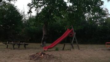 Une aire de jeux avec un toboggan rouge, un banc en bois et un arbre.