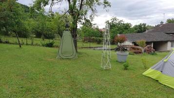 A grassy outdoor area with a tent and a metal structure, likely a tower, in a rural setting with trees and a house in the background.