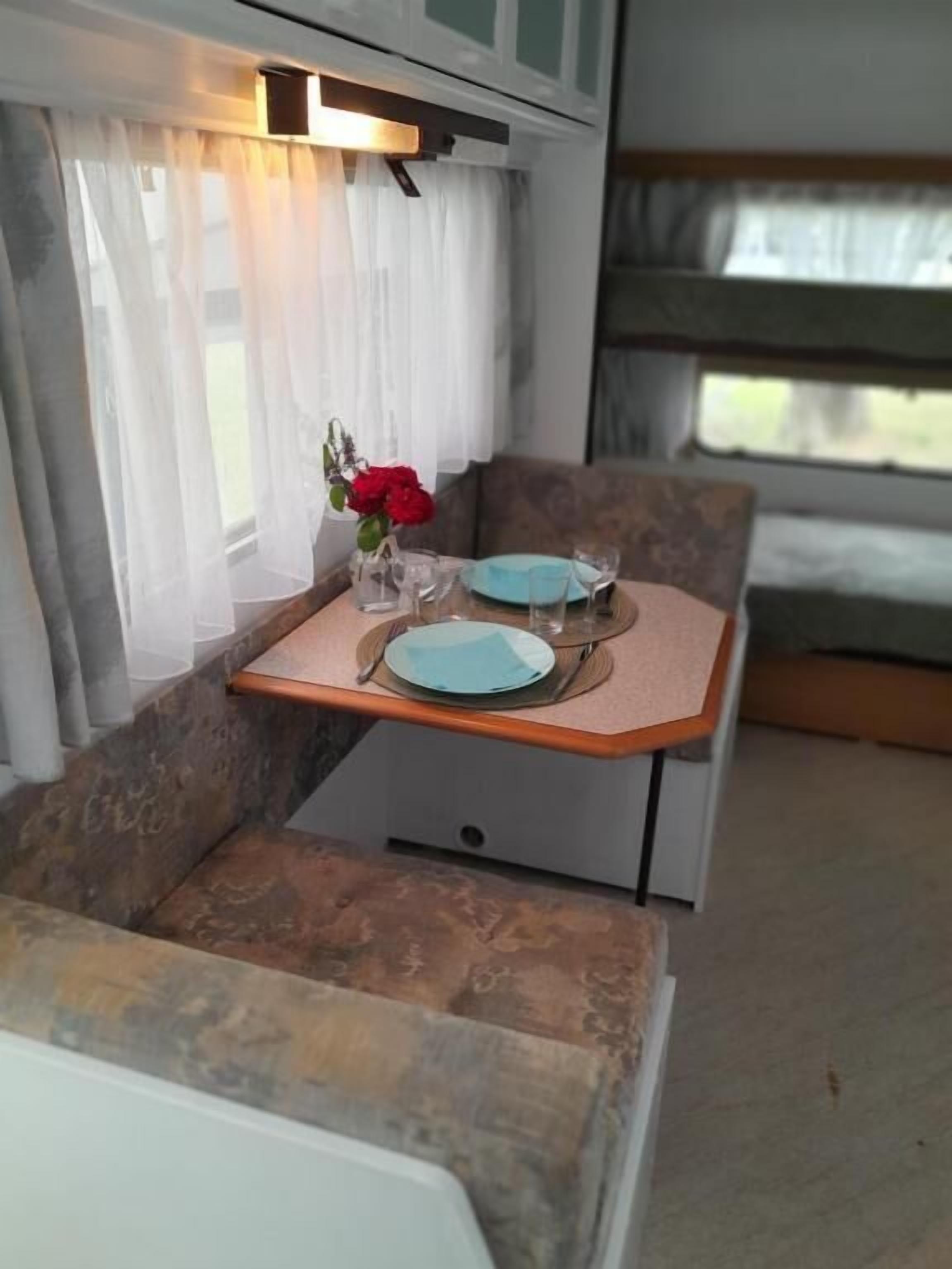 Un comedor pequeño con una mesa dispuesta para dos, un jarrón con flores rojas y una ventana con cortinas traslúcidas.