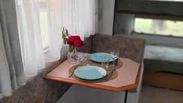 Un comedor pequeño con una mesa dispuesta para dos, un jarrón con flores rojas y una ventana con cortinas traslúcidas.