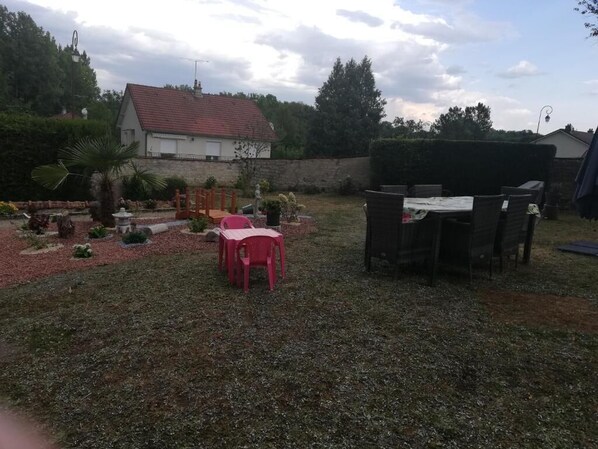A backyard with a red plastic chair, a table, and a barbecue area. - Gîte du capitaine (Maranville)