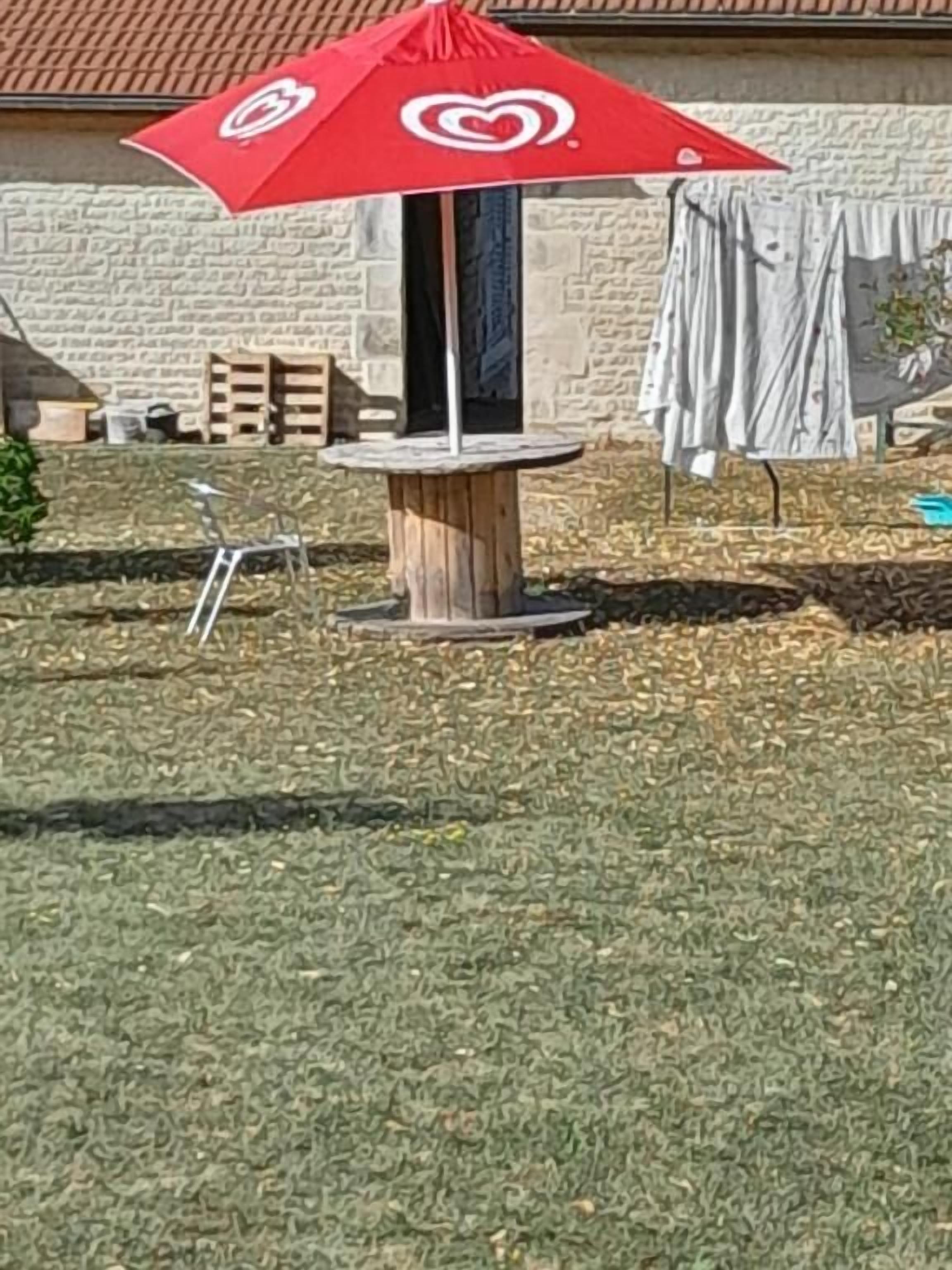 Un espacio patio con una sombrilla roja, una mesa de madera y un muro de piedra al fondo.