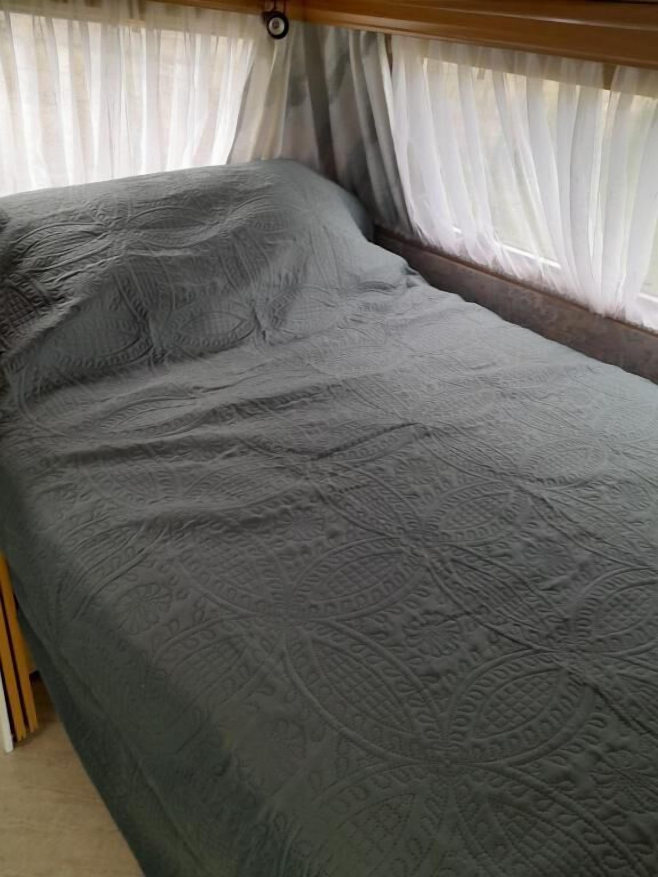 Een bed met een grijs geweven sprei en witte gordijnen.