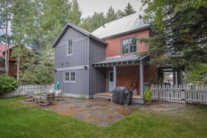 A two-story house with a grey exterior and a brick section, a stone patio, and a white picket fence.