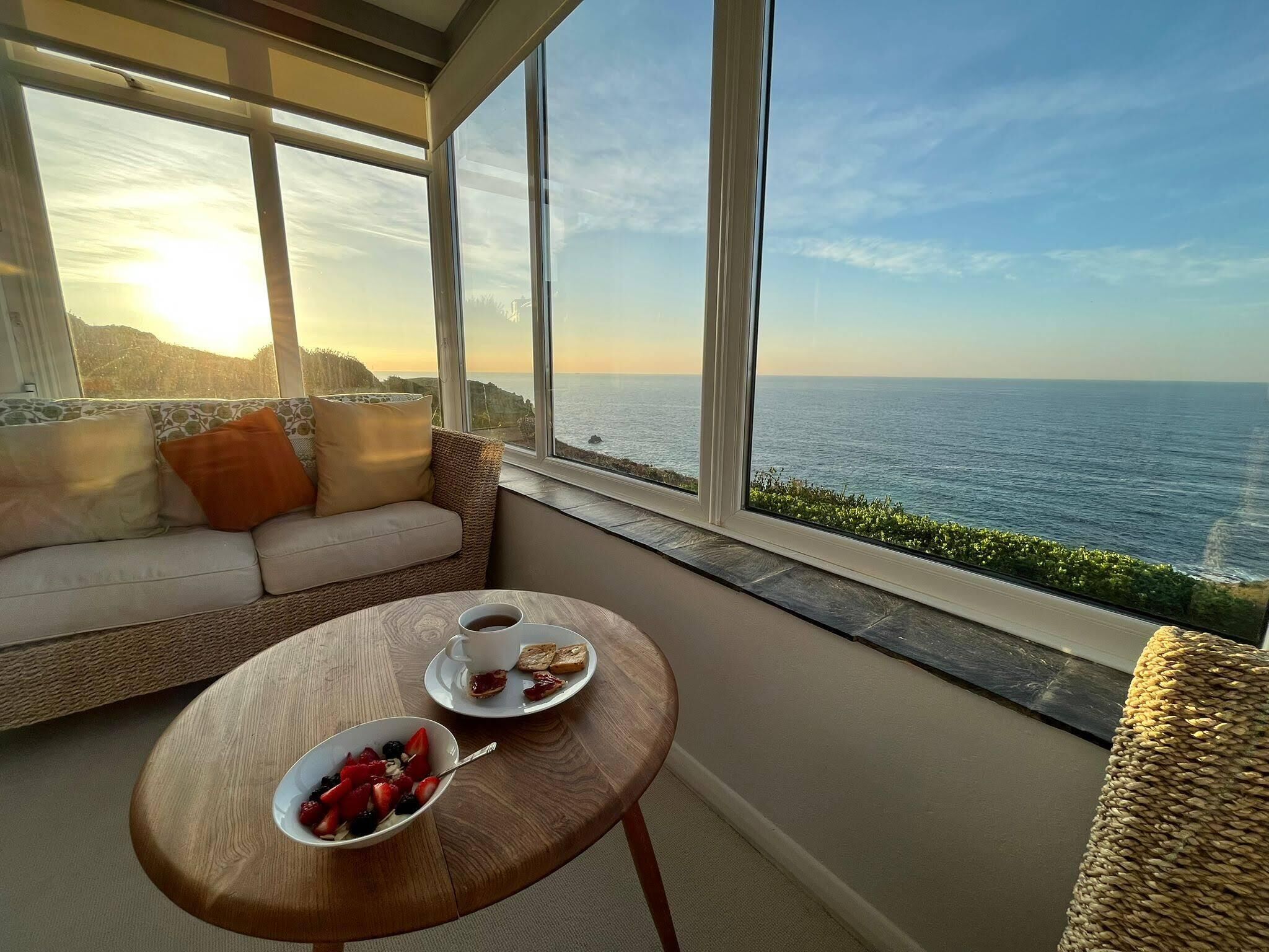 Un balcone con vista sul mare, un divano in vimini, un tavolo in legno con la colazione e un'ampia finestra.