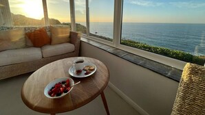 En balkon med havudsigt, en fletsofa, et træbord med morgenmad og et stort vindue.