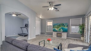 A modern living room with a grey sofa, a glass coffee table, a treadmill, and a large TV screen displaying a nature scene. - Pool! Workout Room, Treadmill, Grill, Pet Friendly! (Peoria)