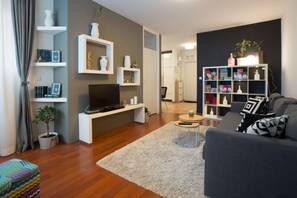 Una sala moderna con un sofá gris, un televisor sobre un mueble blanco, una estantería y una pequeña mesa de centro.