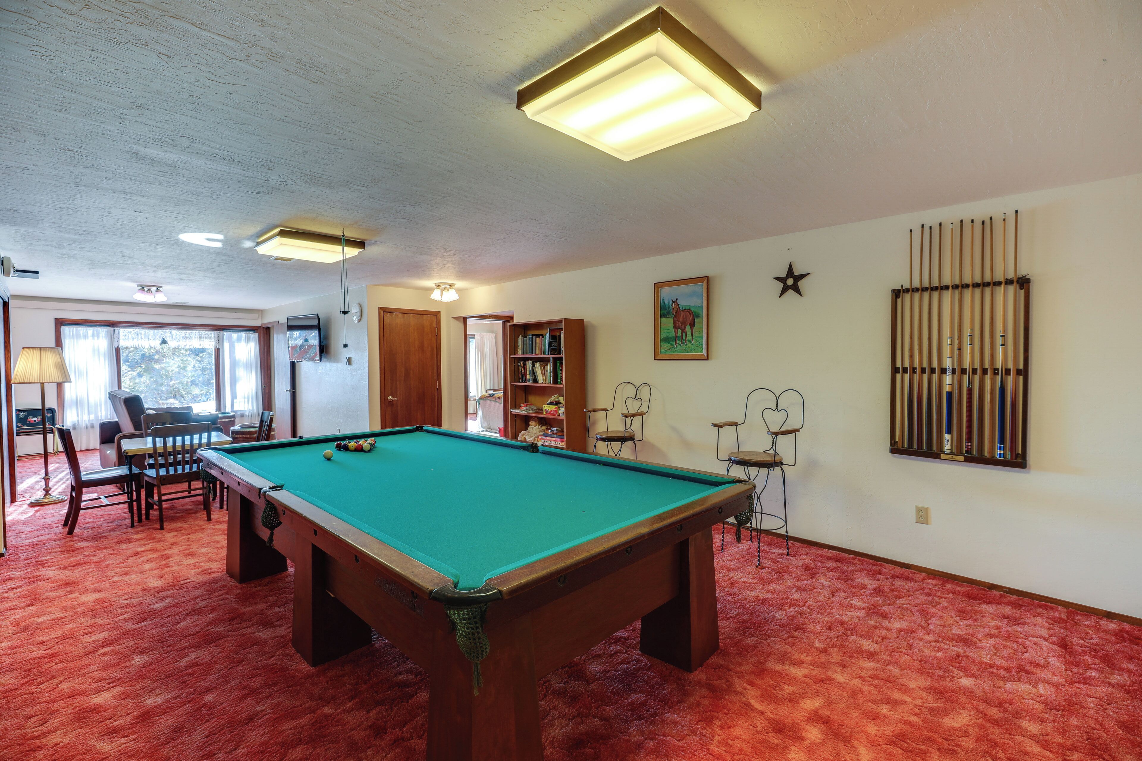 Una mesa de billar en una habitación con alfombra roja, una estantería y un ventanal con cortinas.