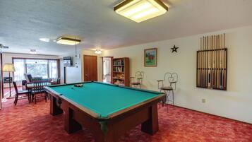 Una mesa de billar en una habitación con alfombra roja, una estantería y un ventanal con cortinas.