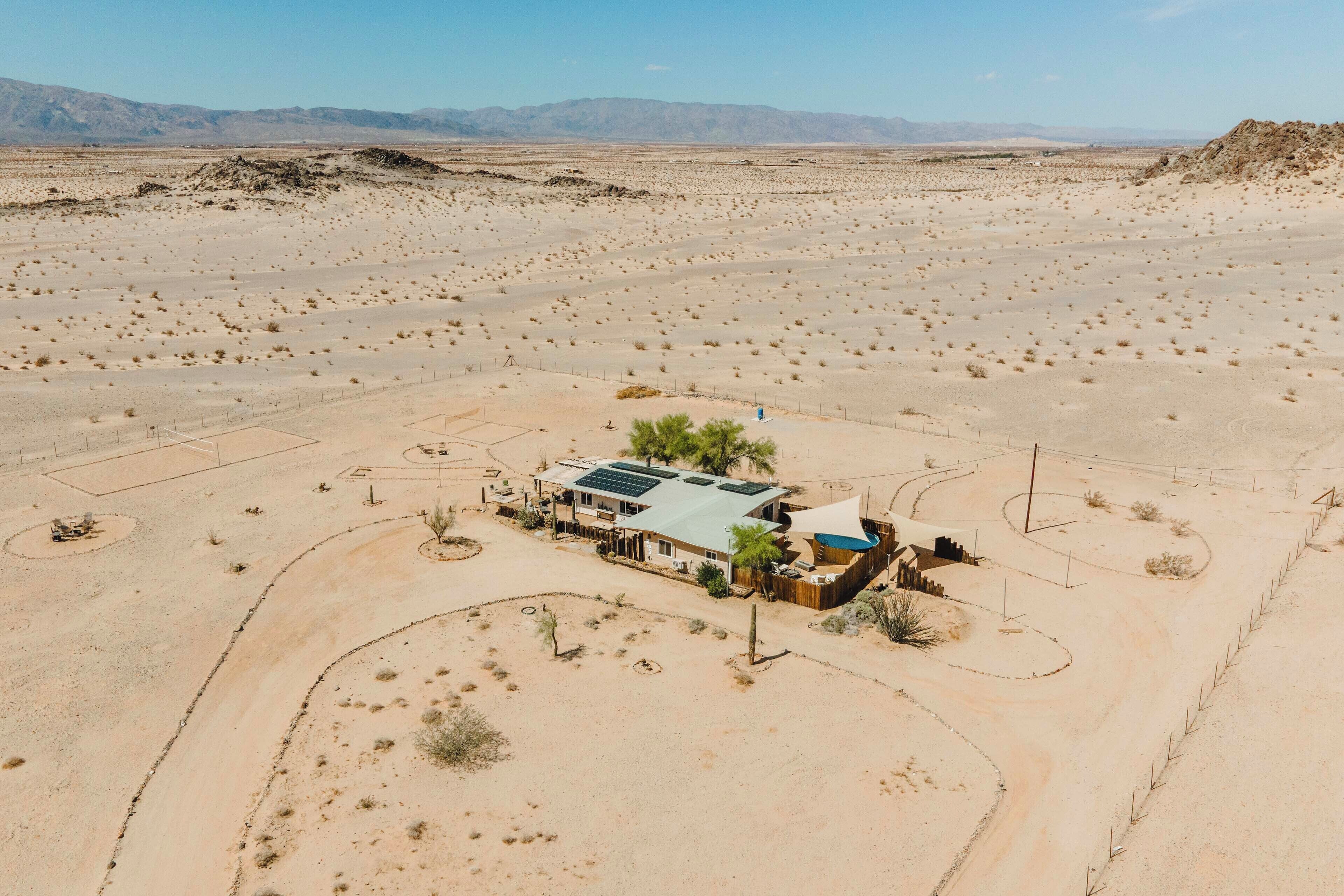 A desert landscape with a single house featuring a solar panel roof, surrounded by sparse vegetation and a fenced area.