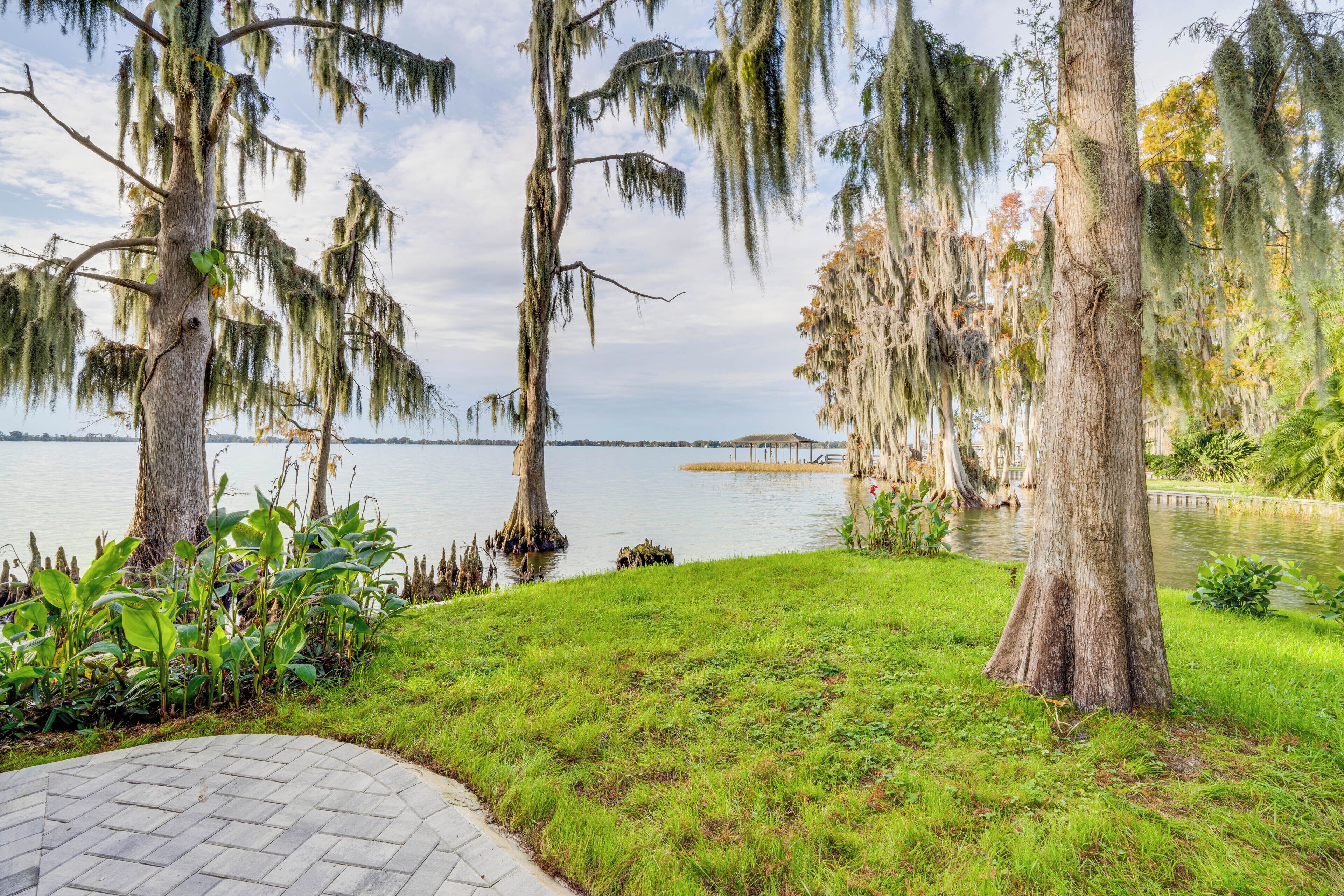 Un parc paisible au bord du lac, avec de grands arbres, une allée pavée et une vue lointaine sur une maison de l’autre côté de l’eau.