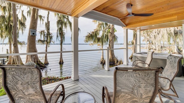 A covered deck with wooden flooring, white railings, and a view of the water. There are chairs and a table, and a ceiling fan is visible. - Hot Tub & Fire Pit: Lakefront Home on Lake Eloise (Winter Haven)