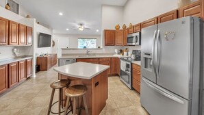 A modern kitchen with wooden cabinets, a stainless steel refrigerator, and a central island with stools. - Summer All Year Round 2 Bedroom Home by RedAwning (Phoenix)