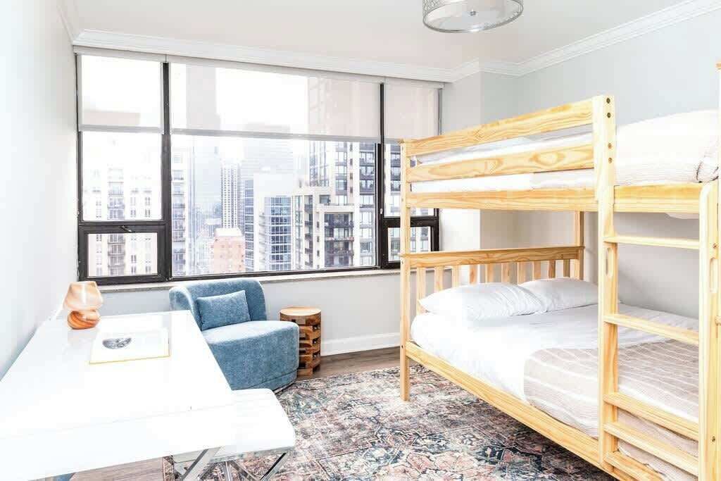 Quarto com beliche, escrivaninha, cadeira e uma grande janela com vista para a cidade.