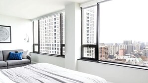 A modern bedroom with a large bed, a gray sofa, and a city view through large windows. - Luxury Designer Penthouse 3802 | Pool | Gold Coast (Chicago)