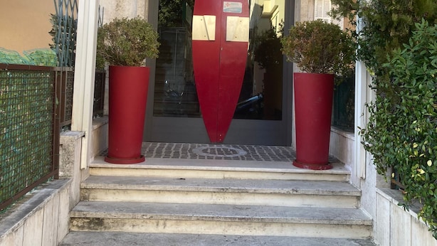 Une entrée d’immeuble avec un panneau triangulaire rouge, des marches menant à l’entrée et deux plantes en pot de chaque côté.