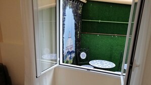 Un petit balcon donnant sur un immeuble, visible à travers une fenêtre, avec un tapis d’herbe artificielle verte et deux petites tables rondes.