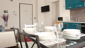 Un comedor con una mesa de vidrio dispuesta para cuatro personas, un sofá blanco, armarios de cocina color turquesa y un televisor de pantalla plana montado en la pared.
