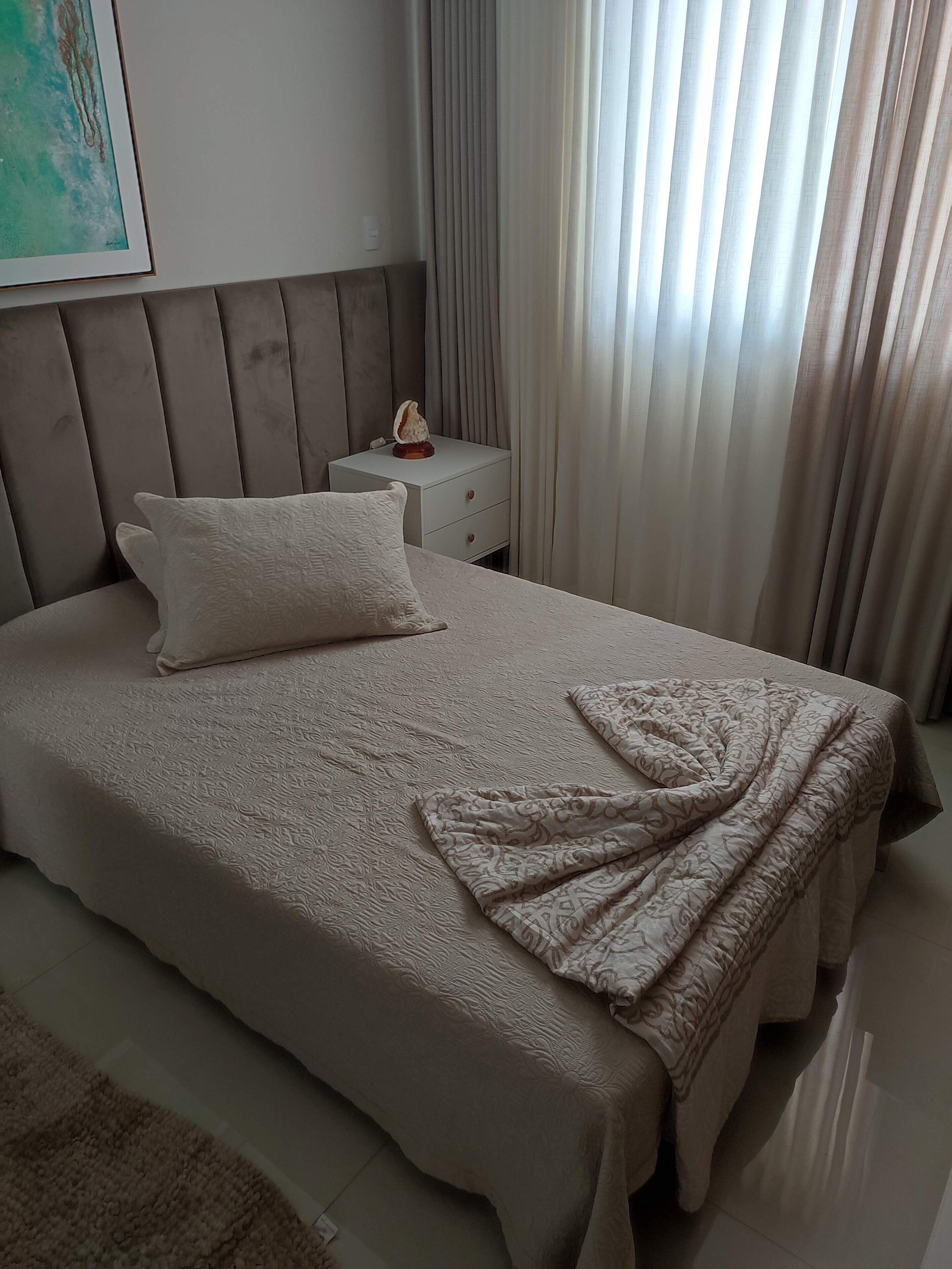 A neatly made bed with a patterned bedspread and a folded blanket, a bedside table with a decorative item, and a window with sheer curtains.