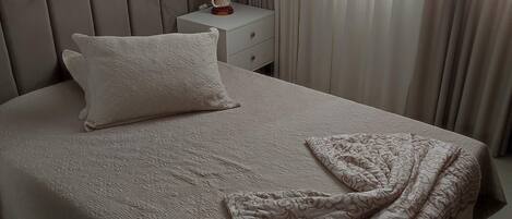 A neatly made bed with a patterned bedspread and a folded blanket, a bedside table with a decorative item, and a window with sheer curtains.