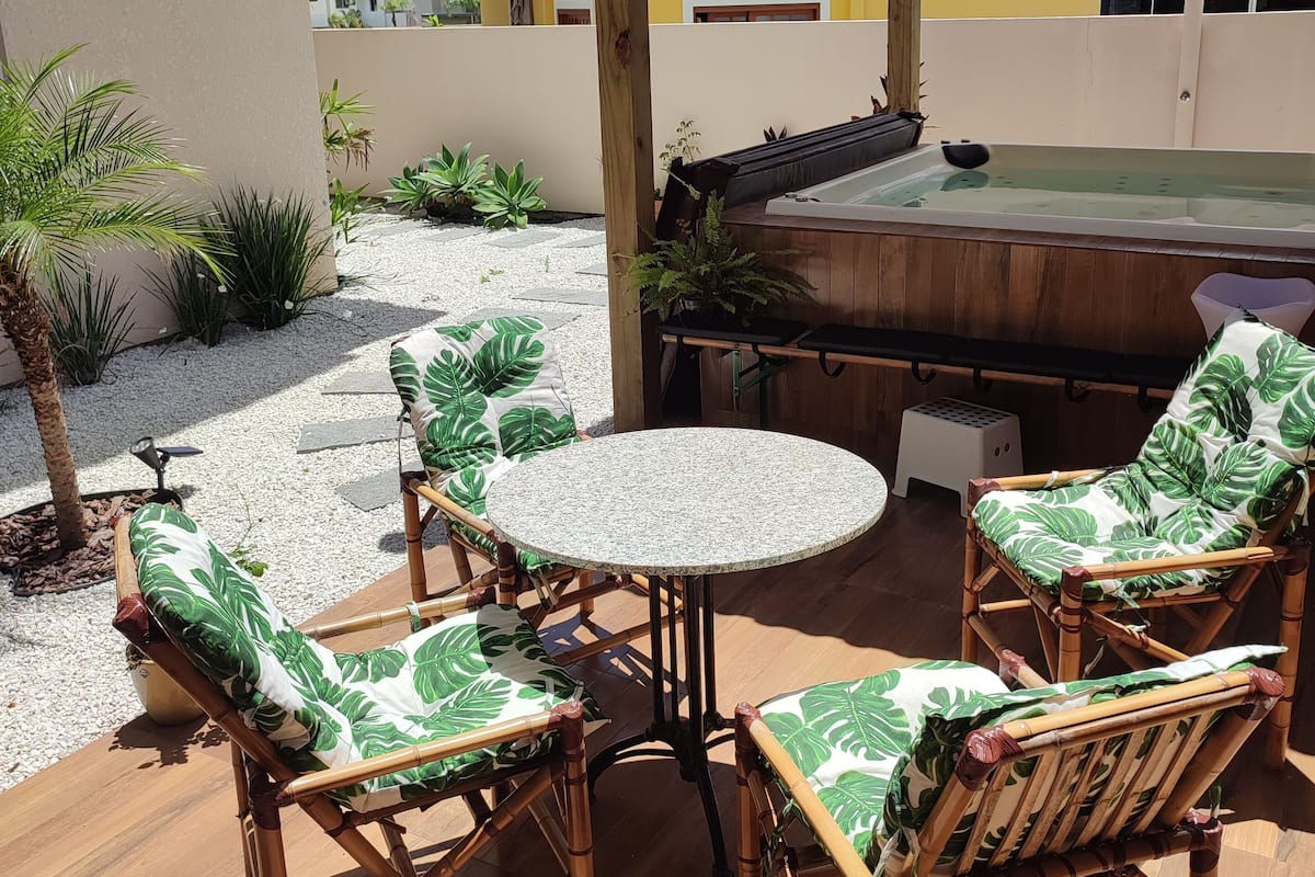 Une terrasse extérieure avec une table ronde et quatre chaises en bambou dotées de coussins verts, un jacuzzi et une pergola.