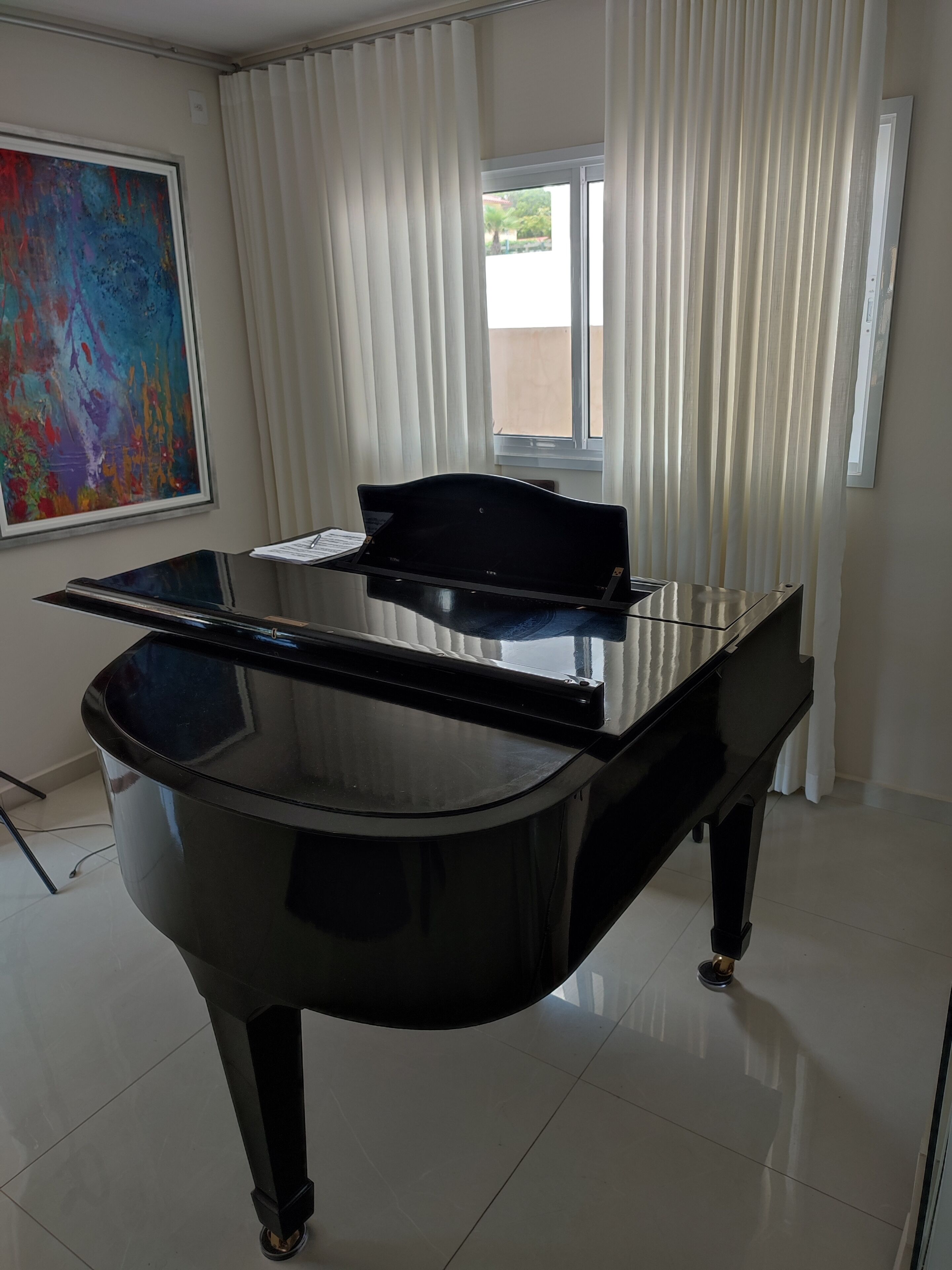 A grand piano in a spacious room with a large window, a colorful abstract painting on the wall, and a clean, tiled floor.