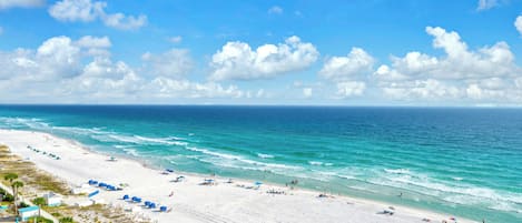 A beach with white sand, clear blue water, and a few umbrellas.