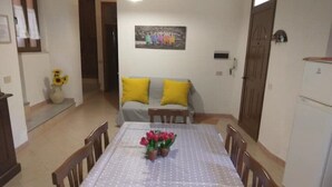 Un coin repas avec une table dressée pour quatre personnes, entourée de chaises en bois. On aperçoit un vase avec des fleurs sur la table, et un canapé avec des coussins jaunes à l’arrière-plan.