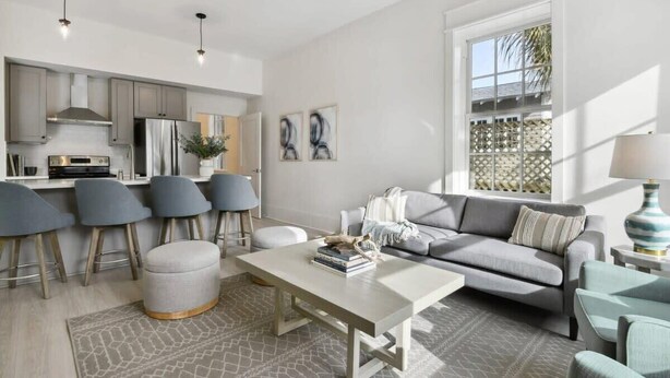 A modern living room with a grey sofa, a coffee table, and a dining area with bar stools. - Bay St Louis Condos, Old Town BSL (Bay St Louis)