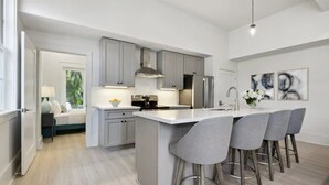 A modern kitchen with grey cabinets, a central island with bar stools, a refrigerator, and a coffee machine. - Bay St Louis Condos, Old Town BSL (Bay St Louis)