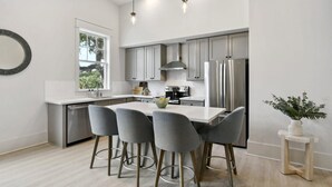 Una cocina moderna con una isla central, taburetes grises, electrodomésticos de acero inoxidable y un ventanal que proporciona luz natural.
