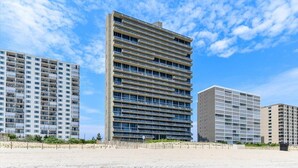 A modern high-rise building with multiple balconies, located on a sandy area with other similar buildings in the background. - Century One-Sunny Oceanfront 2 bed/2 bath with Indoor Pool! (Ocean City)