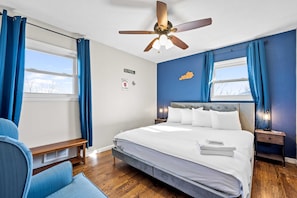 Ein Schlafzimmer mit Bett, Deckenventilator, Nachttisch, einer Bank und einem Fenster mit Vorhängen.